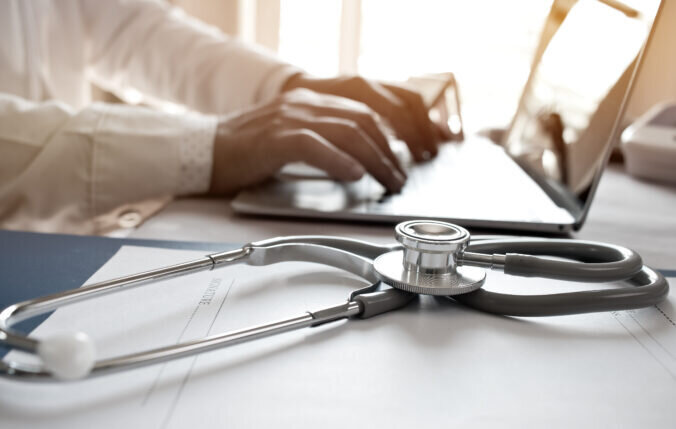 A doctor typing with a stethoscope next to them on the desk