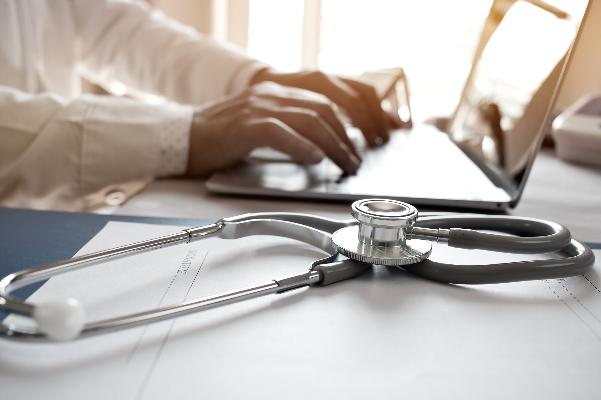 A doctor typing with a stethoscope next to them on the desk